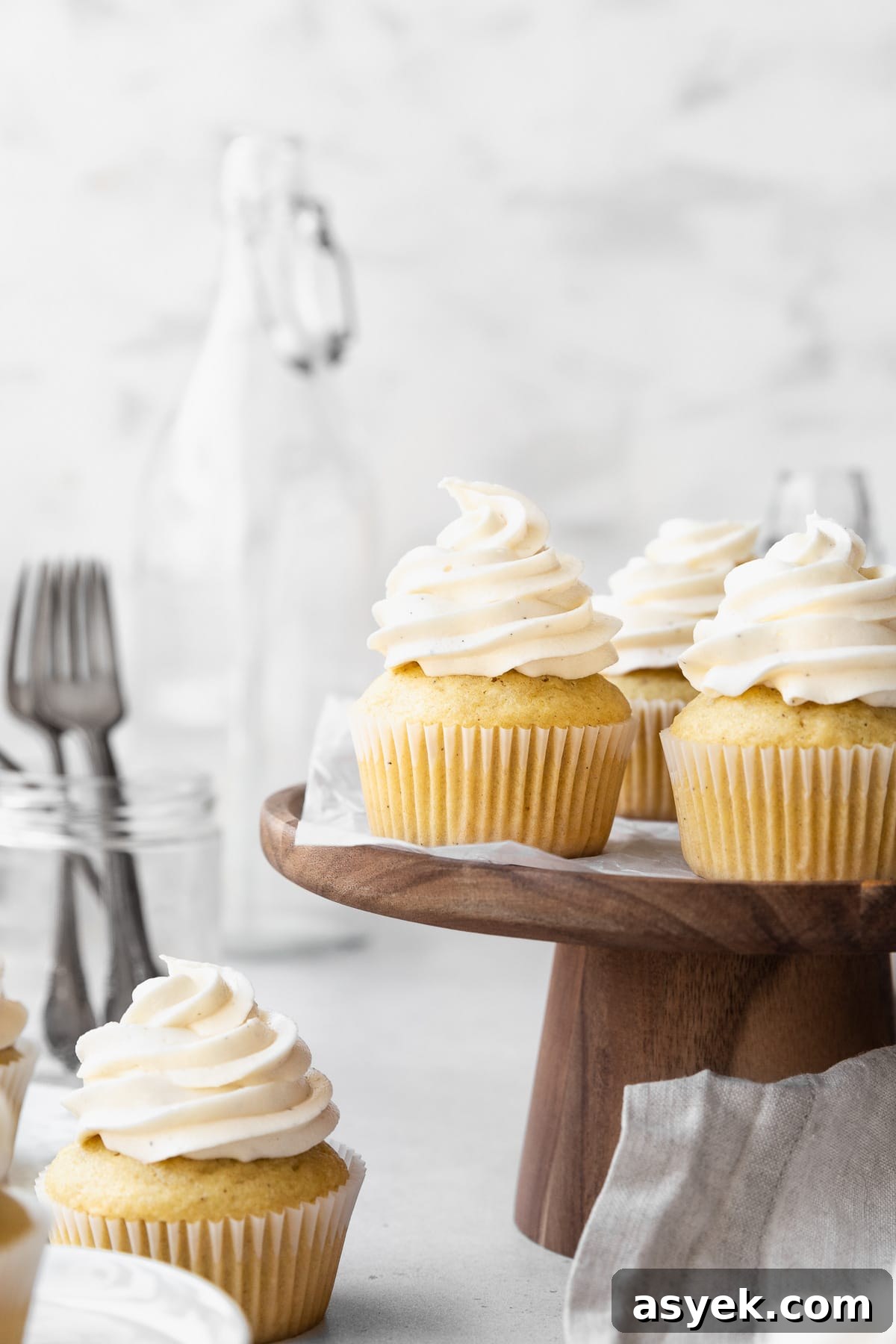 Side on shot of dairy-free vanilla cupcakes on a wooden cake stand with a jar of forks and a large jug in the background.