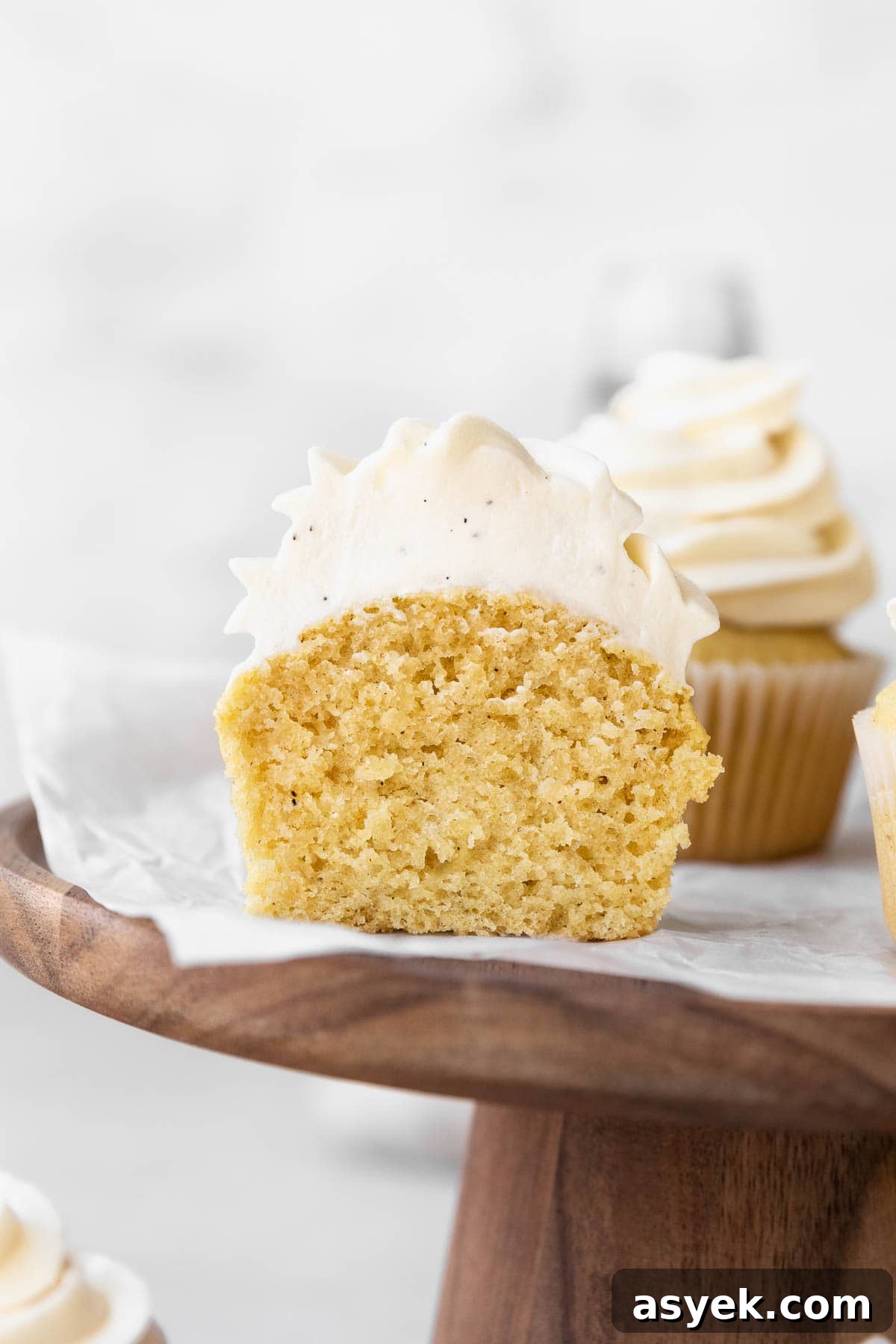 A dairy free vanilla cupcake topped with dairy free vanilla buttercream on a wooden stand, sliced in half to show the fluffy and tender crumb.