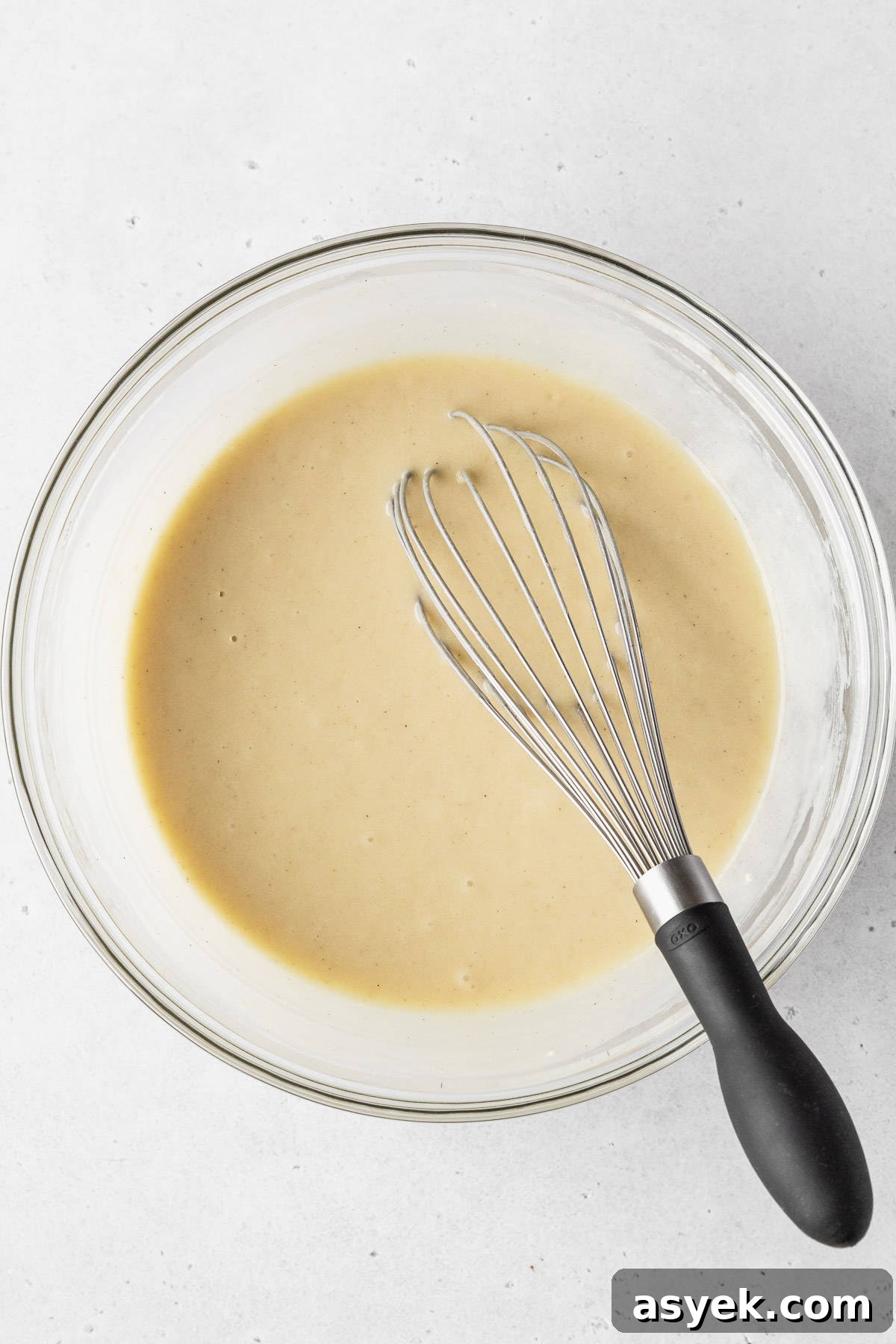 Completed dairy free vanilla cupcake batter in a clear mixing bowl with a black-handled whisk.
