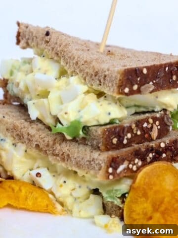 Two halves of an egg salad sandwich stacked, on a plate with sweet potato chips.