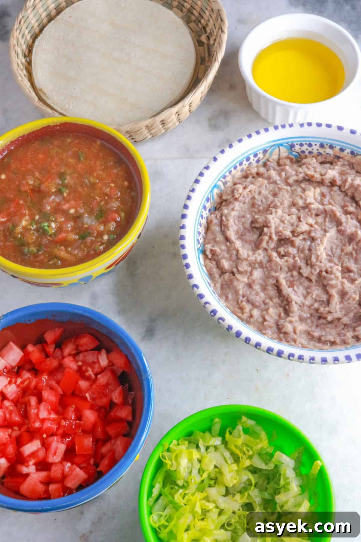 Crispy Mexican Bean Taco Recipe (Tacos Dorados) 3 A basket with corn tortillas, a bowl with oil, a bowl with jalapeño salsa, a bowl with refried beans, another with diced tomatoes and another with shredded lettuce