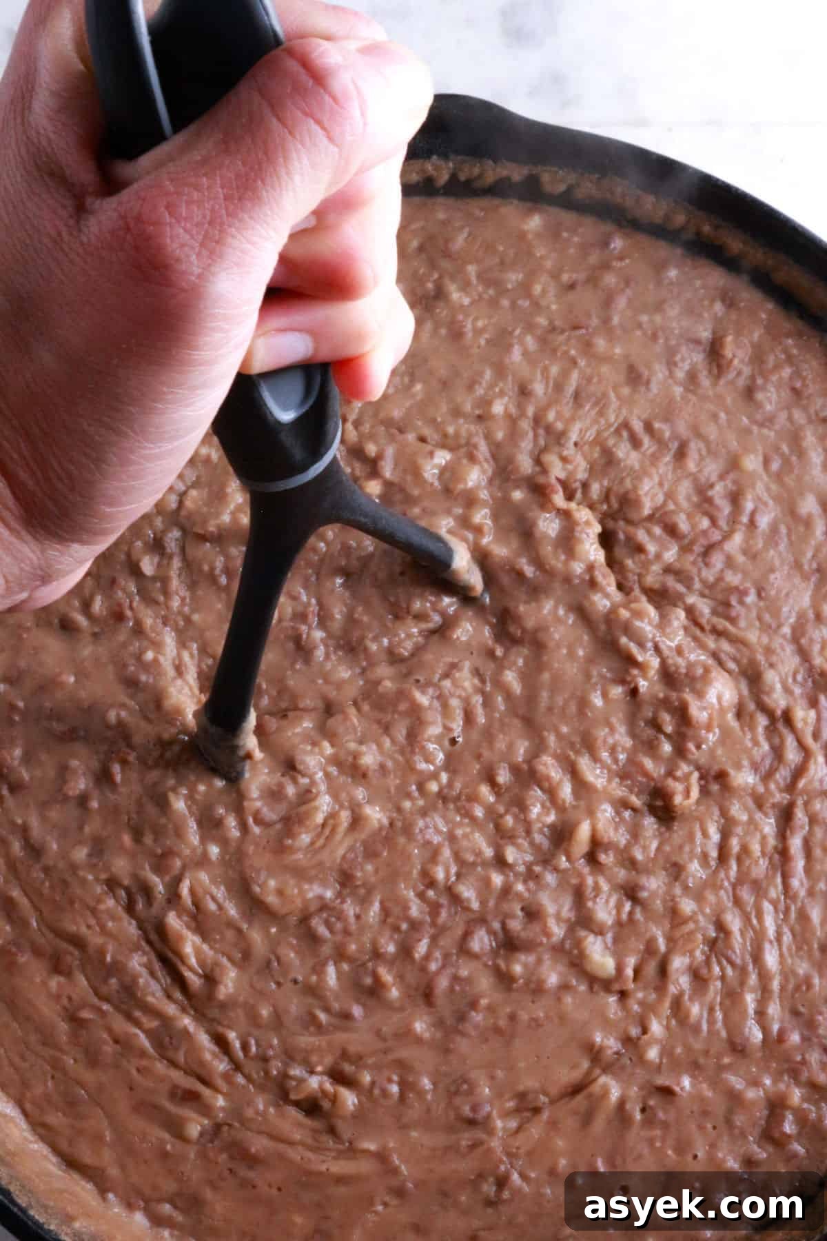 Crispy Mexican Bean Taco Recipe (Tacos Dorados) 4 A hand on a potato masher mashing beans in a cast-iron skillet