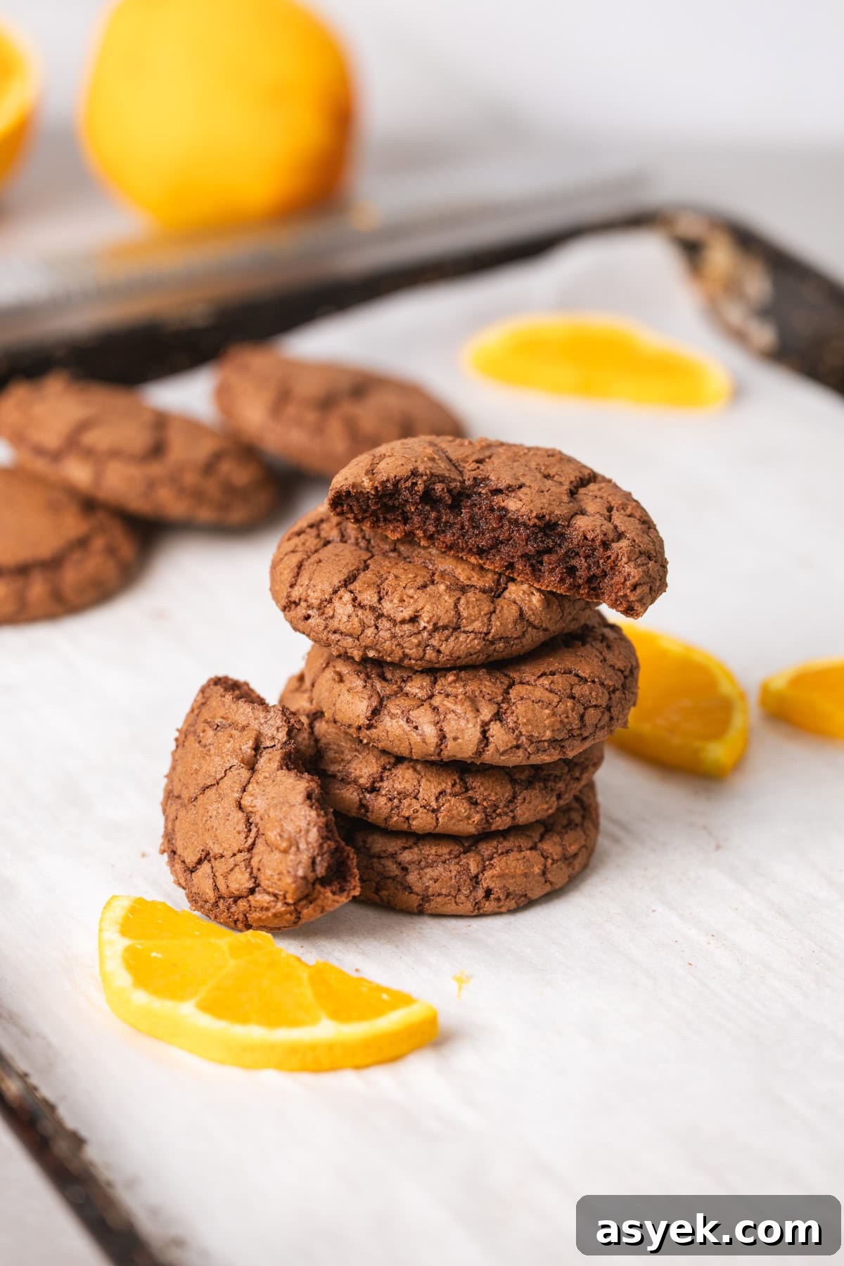 A stack of chewy chocolate and orange cookies on a parchment-lined baking sheet with slices of oranges as a garnish.