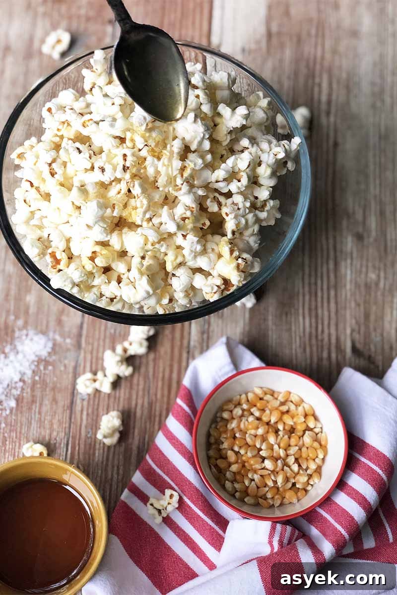 Vertical image of spoon pouring a thick yellow syrup over a bowl of popcorn next to a bowl of the same syrup, a bowl of kernels, and a red-striped white towel on a wooden surface.