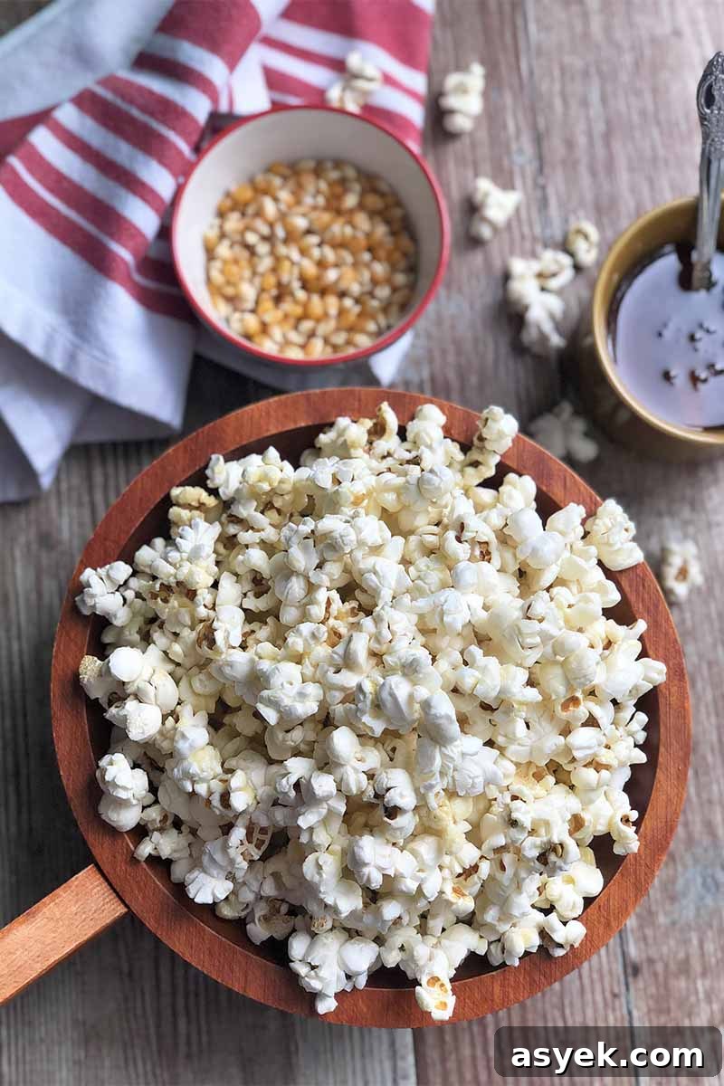 Vertical image of a white towel with red stripes, a cup of honey, a bowl of kernels, and a large wooden bowl filled with popcorn on a wooden surface.
