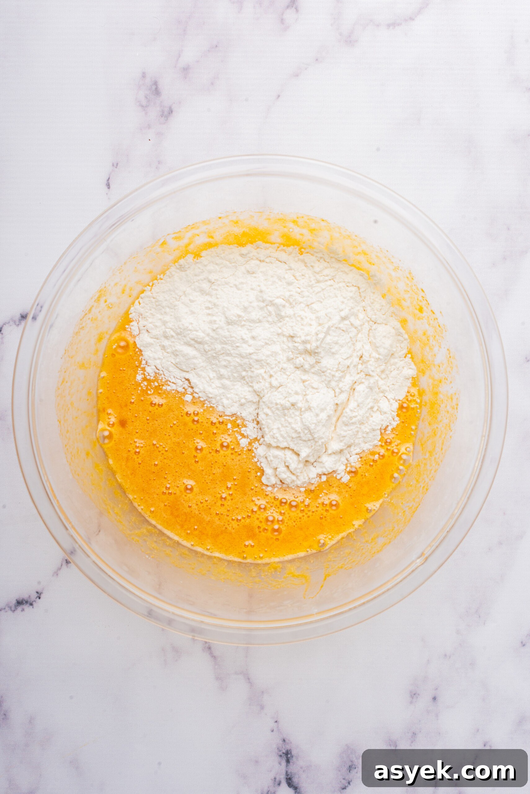 Dry ingredients, including flour, added to the wet mixture in a glass bowl for the Swiss roll cake.
