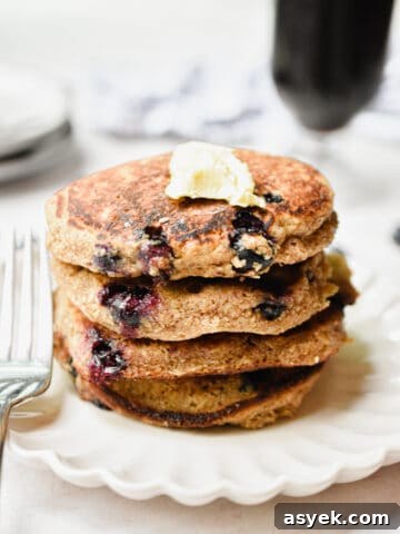 A stack of blueberry oatmeal pancakes have a glass of coffee behind them.