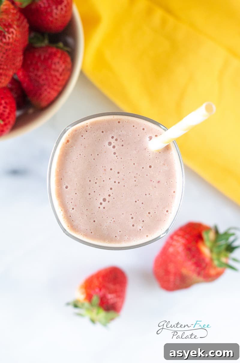 Creamy Strawberry Banana Peanut Butter Smoothie Recipe 6 Top down view of a tall glass filled with strawberry banana peanut butter smoothie, with a yellow and white striped straw. In the background is a bowl of strawberries and a yellow towel.