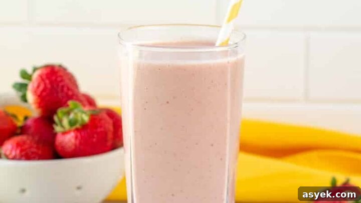 A tall glass filled with strawberry banana peanut butter smoothie, with a yellow and white striped straw. In the background is a bowl of strawberries and a yellow towel.