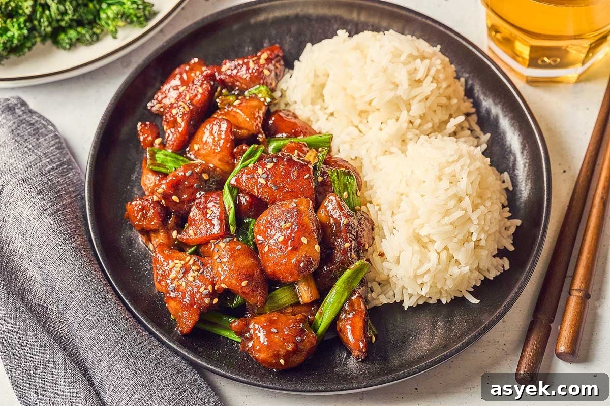A plate featuring Five Spice Chicken Stir Fry with glazed chicken, green onions, and sesame seeds sits invitingly beside two servings of white rice. Chopsticks are elegantly placed alongside a glass of amber liquid, completing the scene.