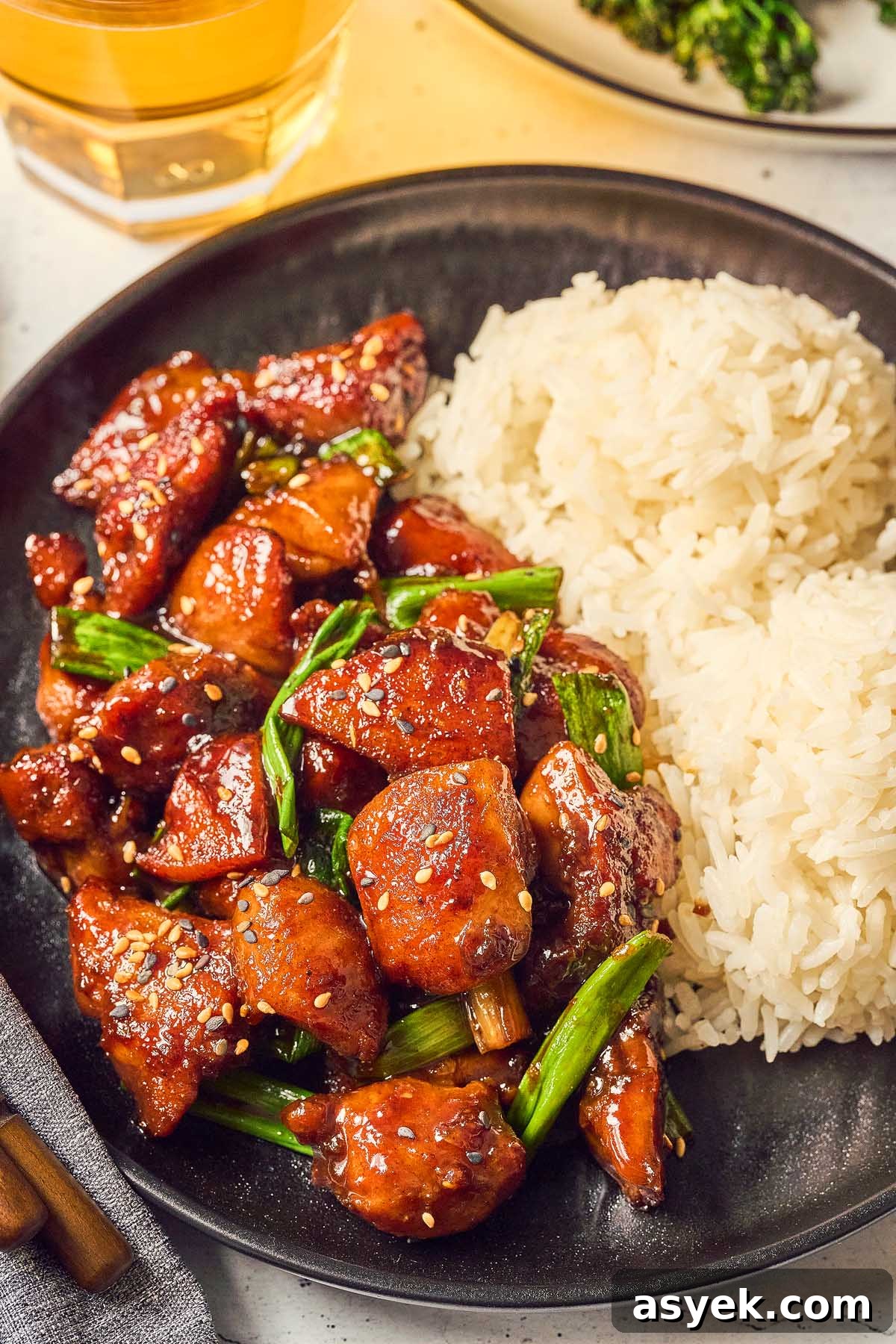 A plate with Five Spice Chicken Stir Fry, featuring glazed chicken pieces, green onions, and a serving of white rice. In the background, a glass of yellow liquid is partially visible.