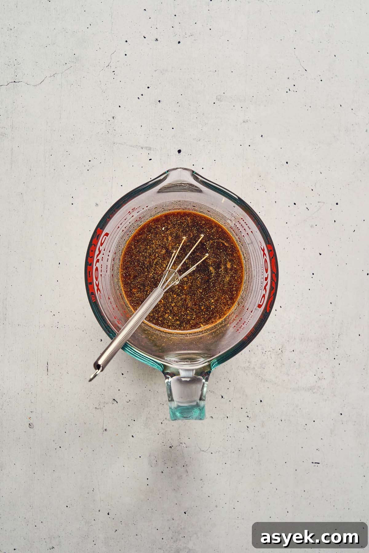 A glass measuring cup filled with a dark liquid, reminiscent of Five Spice Chicken Stir Fry marinade, and a whisk resting on a white speckled surface.