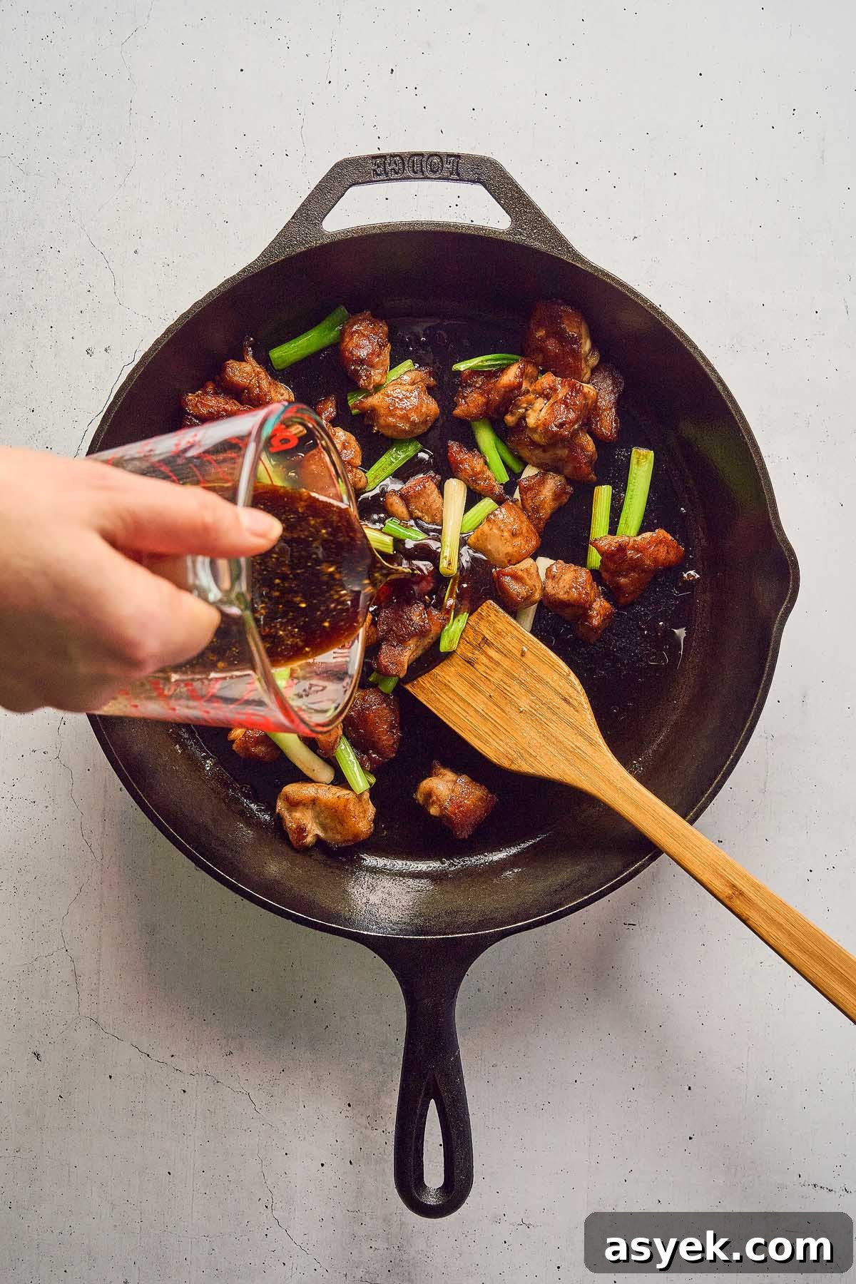 A hand pours sauce from a measuring cup onto Five Spice Chicken Stir Fry, mingling with green onions in a cast iron skillet, as a wooden spatula rests inside.