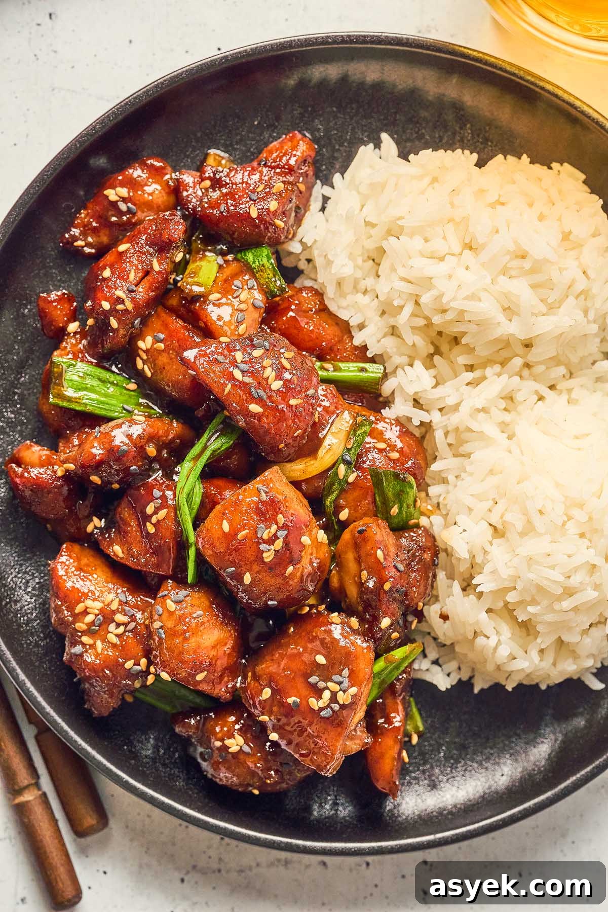 A black plate features Five Spice Chicken Stir Fry, with glazed chicken pieces garnished with sesame seeds and green onions, accompanied by two servings of white rice.