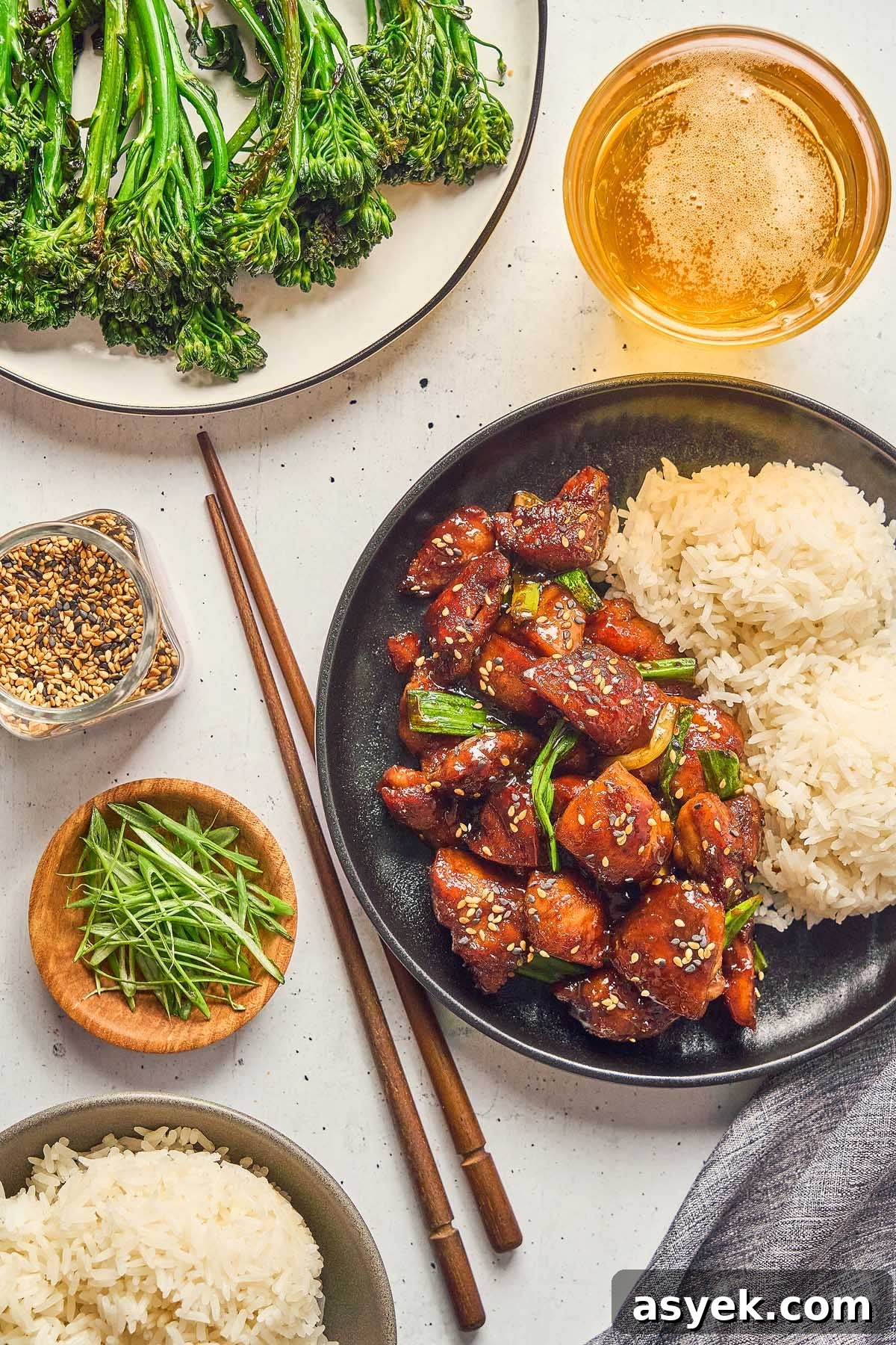 A plate of Five Spice Chicken Stir Fry with rice and green vegetables. Additional dishes feature greens, chopped herbs, and mixed seeds. A glass of a light beverage sits beside the meal on a white surface.