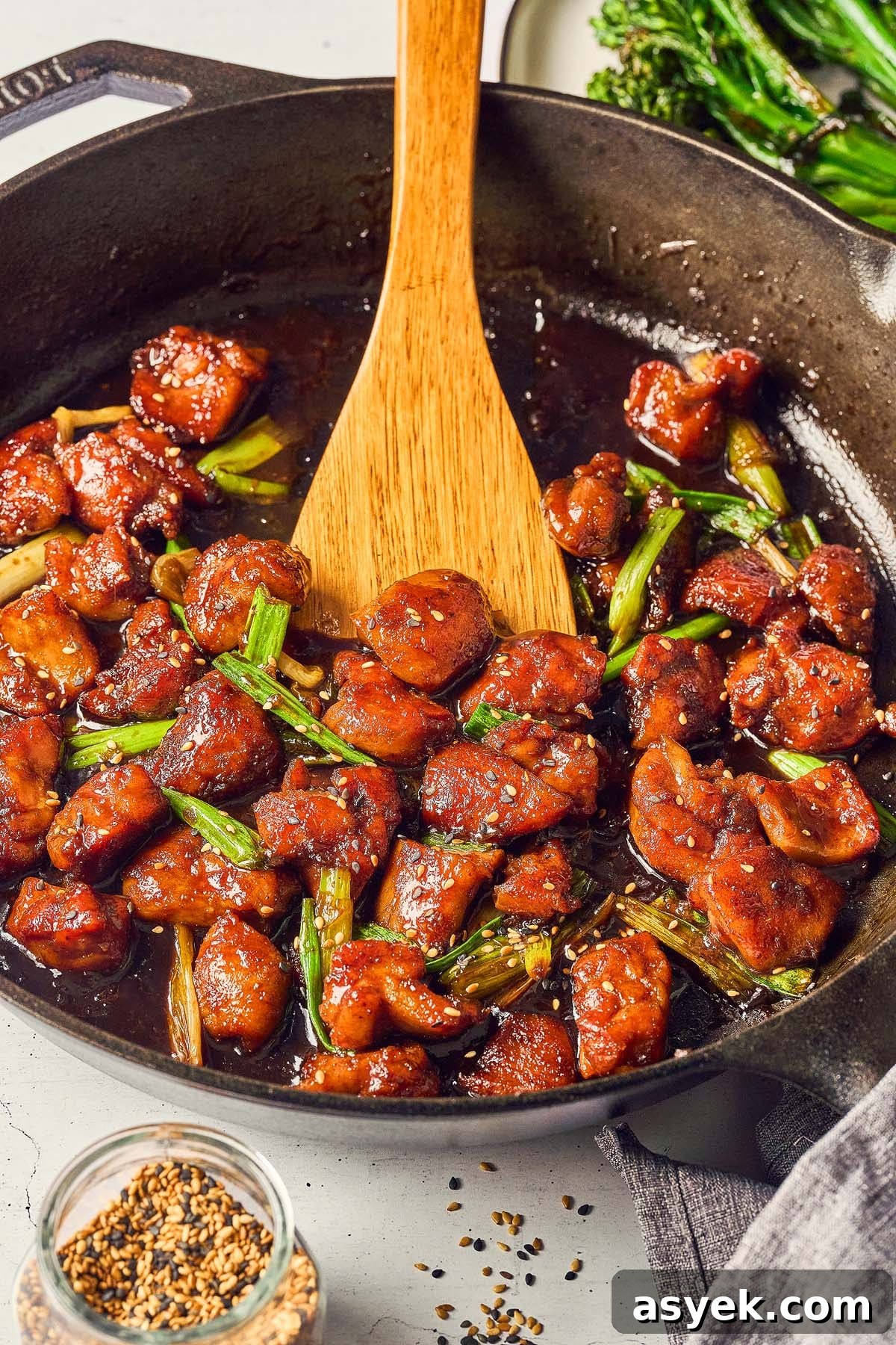 A skillet with Five Spice Chicken Stir Fry, featuring glazed chicken pieces and sliced green onions, alongside a wooden spoon. Nearby, a jar of sesame seeds awaits, with vibrant broccolini adding a touch of green.