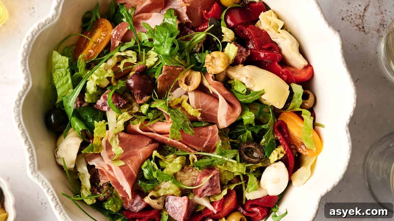 A bowl of salad with mixed greens, prosciutto, cherry tomatoes, olives, artichokes, and roasted red peppers.