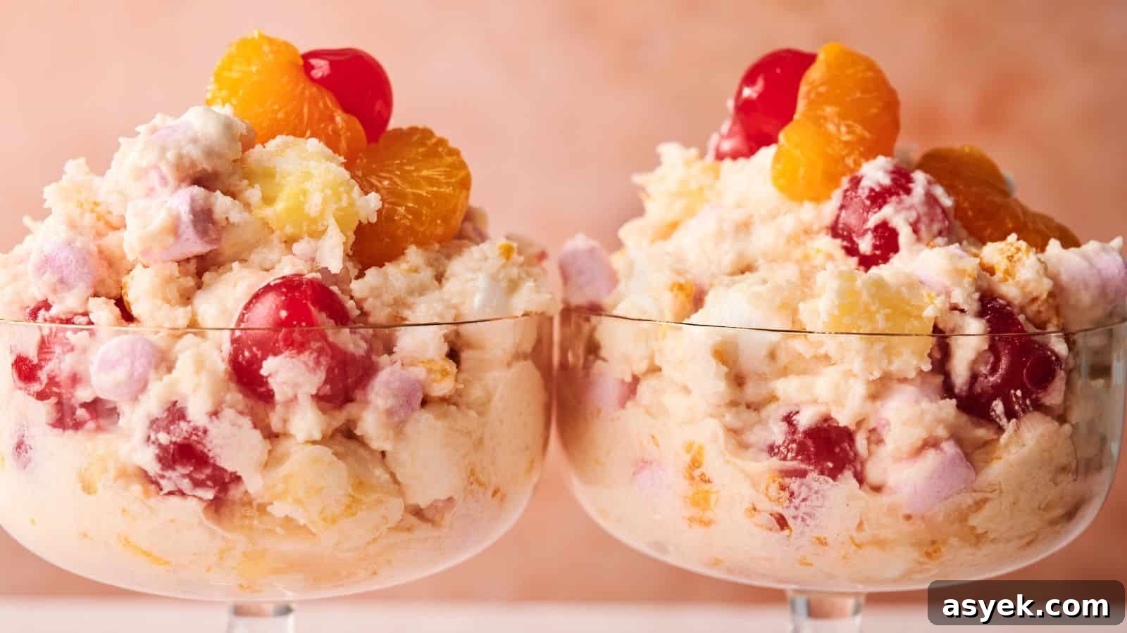 Two glass dessert cups filled with ambrosia salad, featuring whipped cream, cherries, pineapple, mandarin oranges, and marshmallows against a soft pink background.