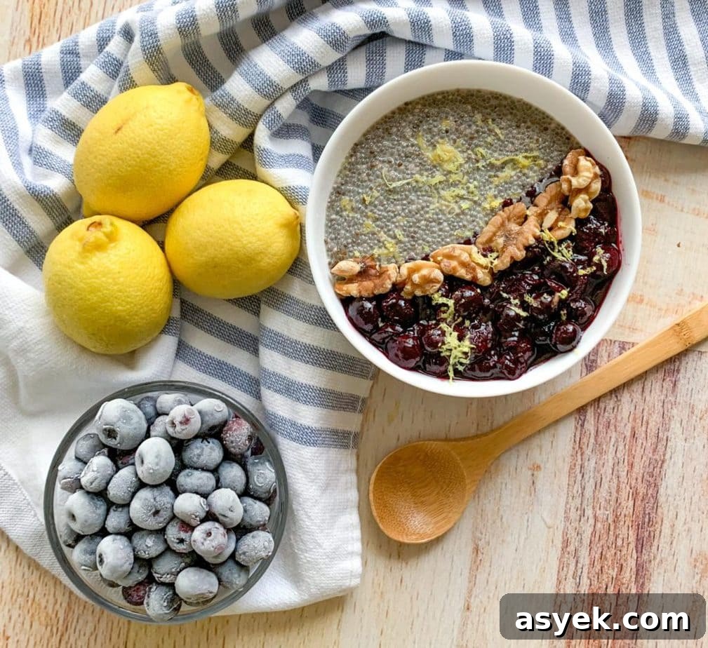 A bowl of chia pudding, topped with blueberry jam and walnuts. A blue striped towel, lemons, blueberries and a wooden spoon alongside.