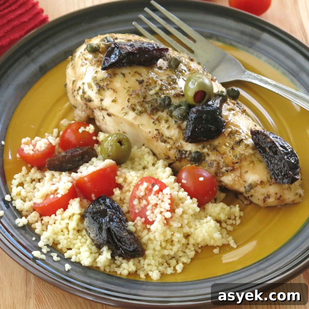 A plate with chicken marbella made with boneless chicken breasts that is topped with prunes, olives, tomatoes, with a side of couscous next to it.