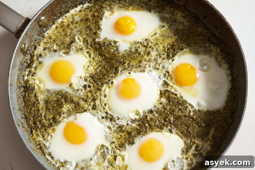 Six cooked sunny-side up eggs in green shakshuka sauce in a large skillet.