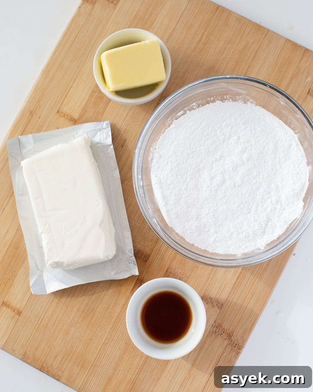 Ingredients for cream cheese frosting on a wooden cutting board. A block of cream cheese, a small white bowl of vanilla extract, a clear glass bowl with fine sugar, and a small clear white bowl with butter. 