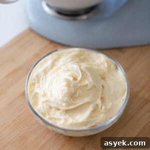 Finished cream cheese frosting in a small round glass bowl on a wooden cutting board with a light blue stand mixer in the background.