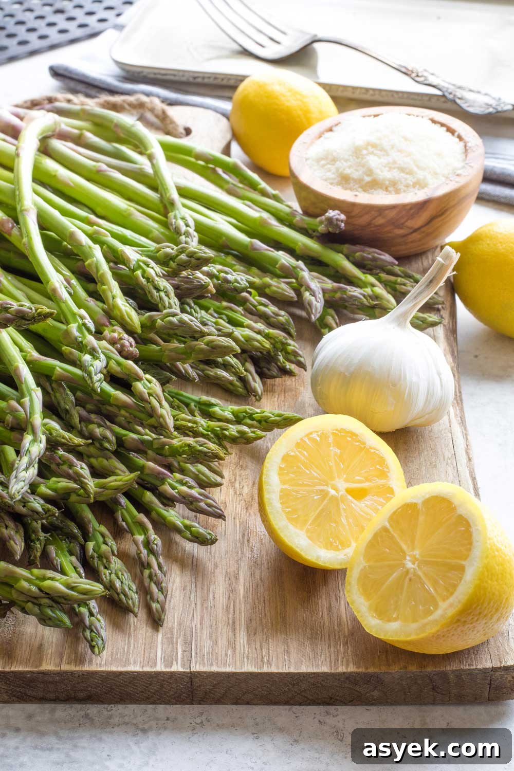 Grilled Asparagus Recipe with Parmesan, Lemon and Garlic 3 Raw asparagus on cutting board with lemons, head of garlic and bowl of parmesan cheese.