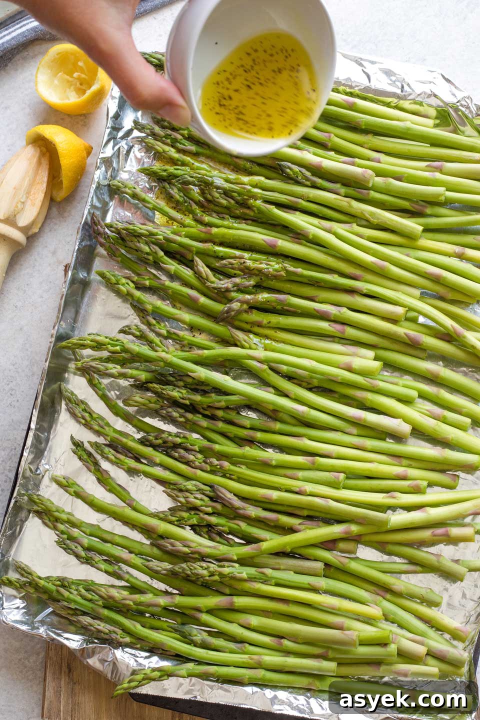 Grilled Asparagus Recipe with Parmesan, Lemon and Garlic 6 Hand pouring marinade on asparagus spread on foil-lined grill pan.