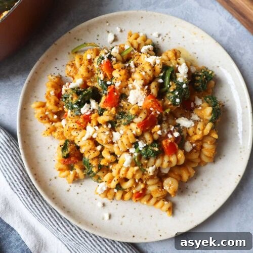 white plate with pasta tossed in a pumpkin feta sauce topped with red bell pepper and spinach