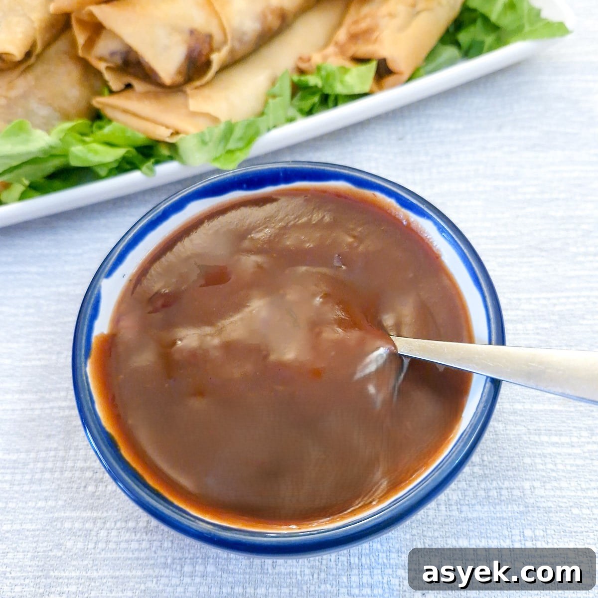 A small bowl of sweet and sour dipping sauce with a spoon. There is a plate of spring rolls in the background.