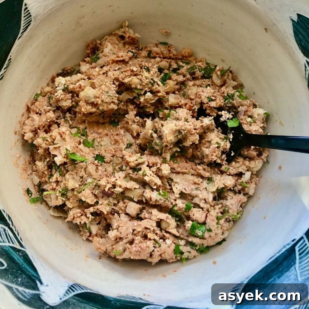 Beige bowl with large black fork mashing together beans, tomato paste, fresh parsley and spices to form vegan meatballs.