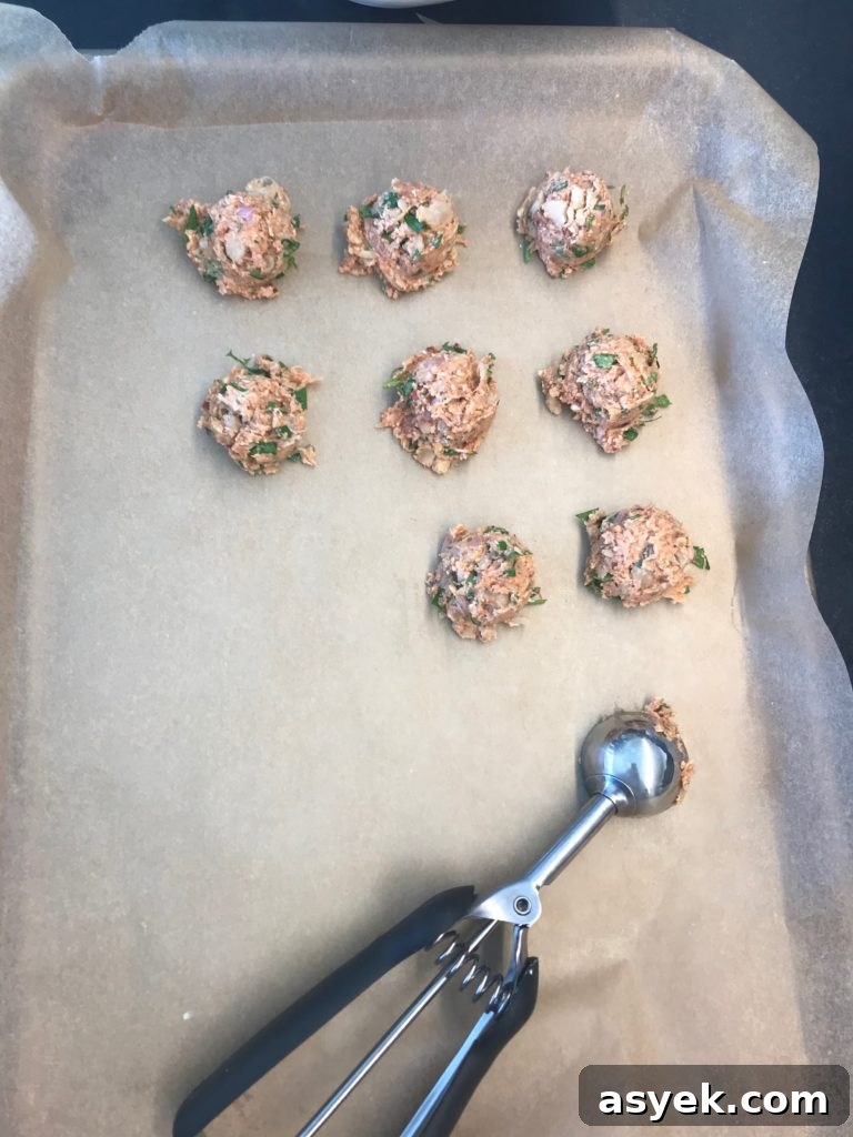Large rimmed baking tray lined with unbleached parchment paper. Small scoop portioning out vegan meatballs in neat rows on prepared tray.