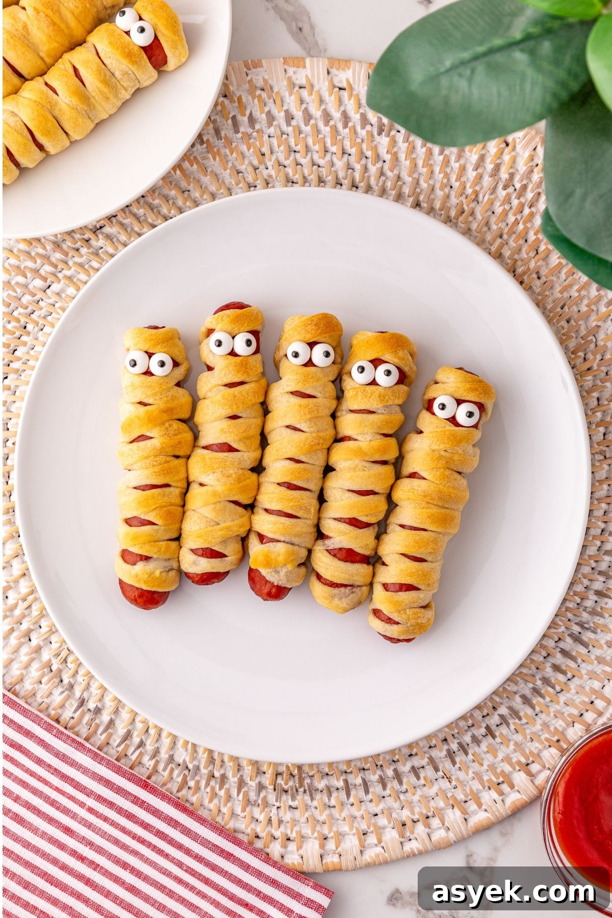 Halloween Mummy Hot Dogs Recipe 2 Mummy hot dogs on a white plate, with candy eyes.
