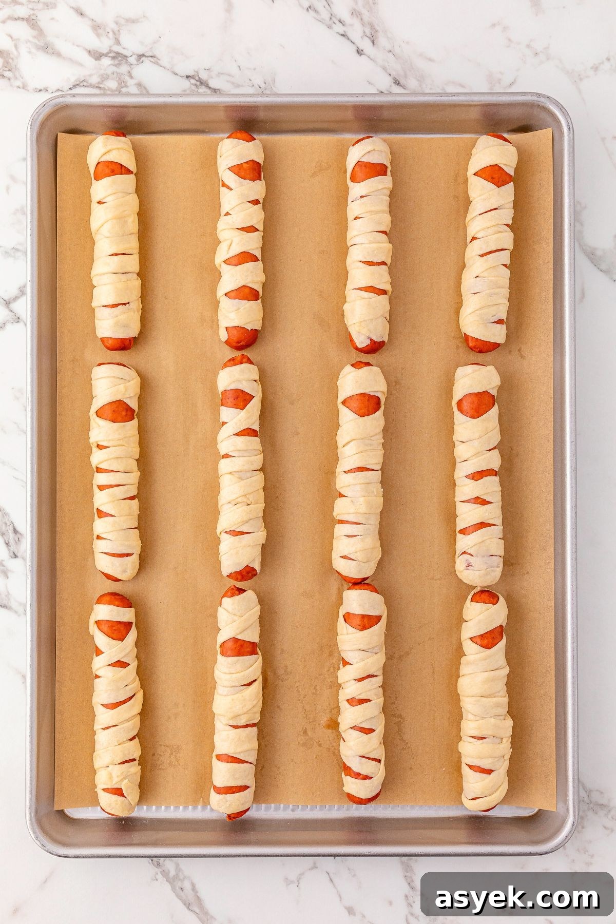 Halloween Mummy Hot Dogs Recipe 5 Uncooked hot dogs wrapped with strips of crescent dough, placed on a baking sheet lined with parchment paper.
