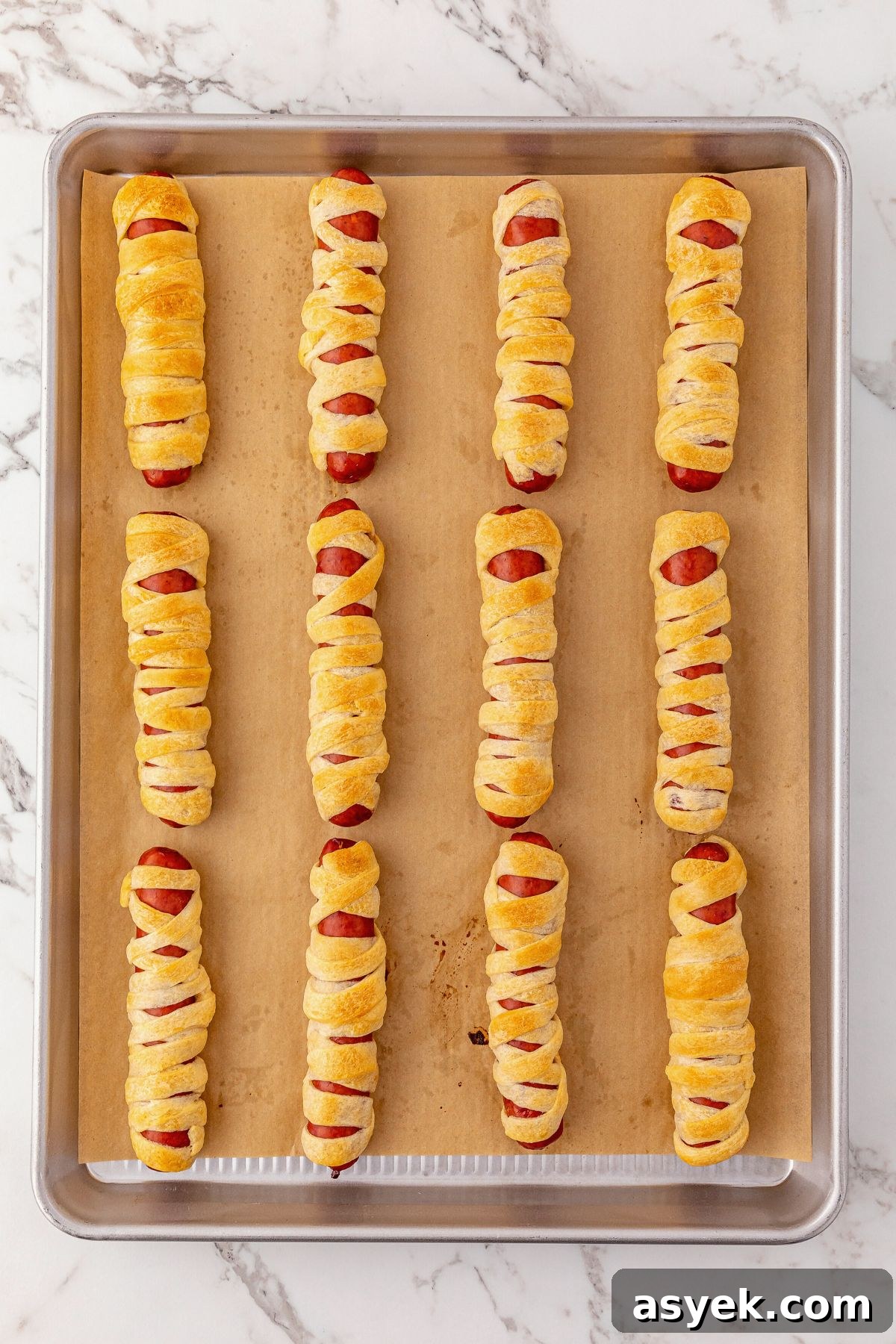 Halloween Mummy Hot Dogs Recipe 6 Hot dogs wrapped in strips of dough and baked until golden brown on a baking sheet, lined with parchment paper.