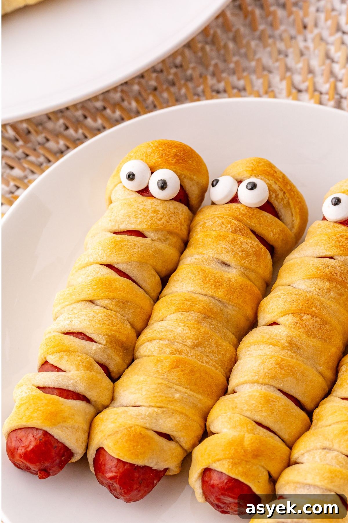 Halloween Mummy Hot Dogs Recipe 8 Mummy hot dogs on a white plate, with candy eyes.