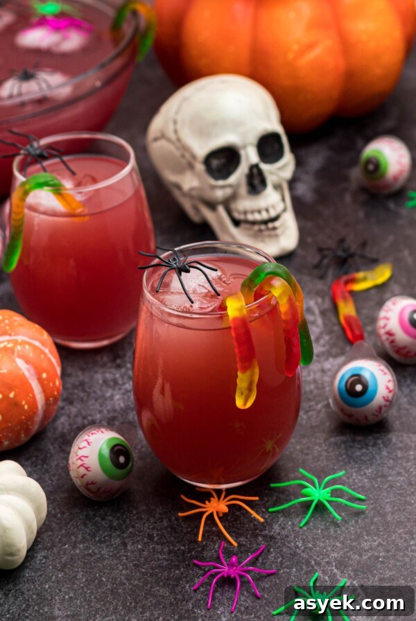 Glass of Halloween Punch in with gummy worms hanging over the side of the glass, in front of plastic skull and spiders.