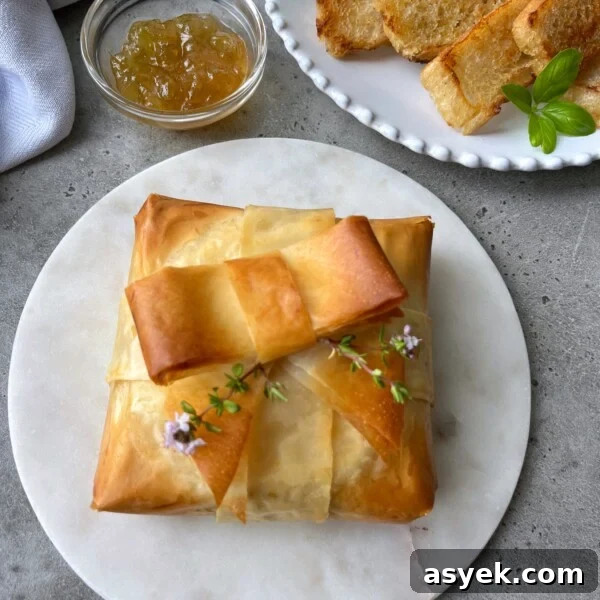 A golden-brown phyllo baked brie, wrapped like a present and garnished with fresh herbs, sits on a marble plate. Nearby are a bowl of jelly and slices of toasted bread on a white plate.