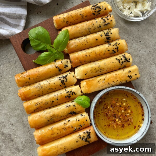 Golden, crispy Phyllo Feta Fries rolled and topped with black sesame seeds are arranged on a wooden board with basil leaves. A small bowl of seasoned olive oil and a bowl of crumbled cheese are nearby.