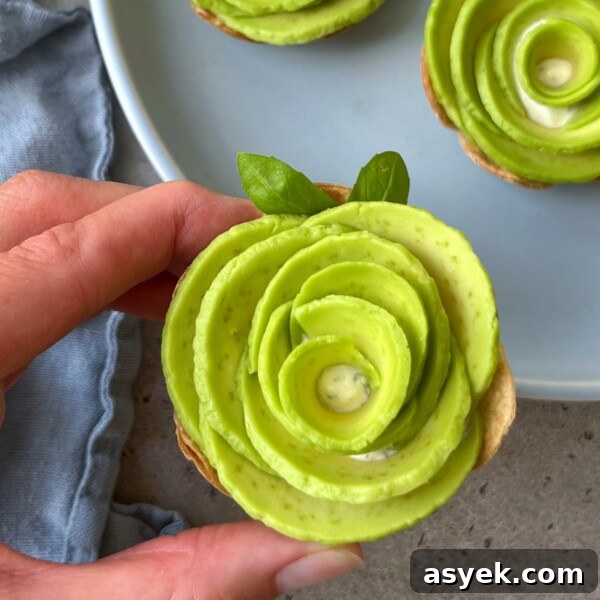 A hand holds a baked tortilla appetizer topped with avocado slices arranged in a rose shape, garnished with basil leaves. This dish sits before a blue napkin and a plate with more avocado bites in the background.