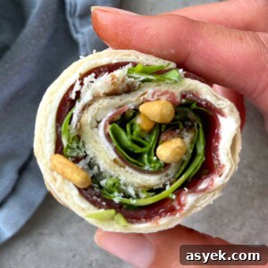 A hand holds a pinwheel-shaped wrap filled with leafy greens, cream cheese, fruit preserves, and a few nuts, making for one of those tasty health bites rolled in a tortilla.