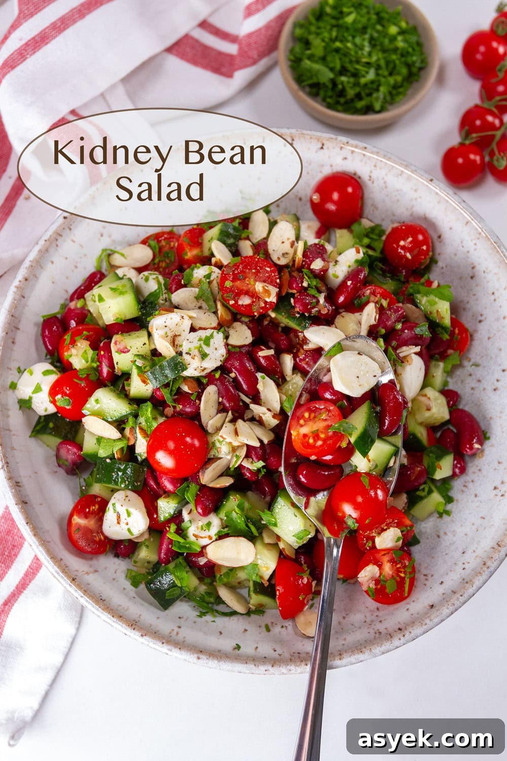 Zesty Kidney Bean Salad with Lemon and Fresh Herbs 2 A white bowl filled with a kidney bean salad with a serving spoon, parsley and cherry tomatoes nearby