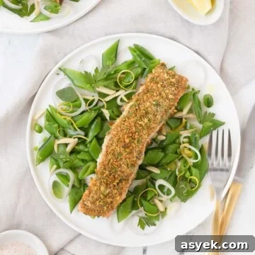 A pistachio crusted salmon filet on a bed of bright green sugar snap peas with a fork.