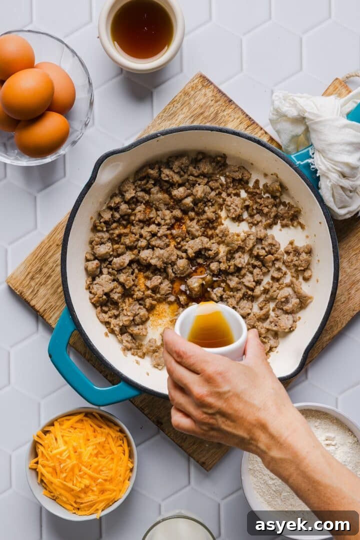 Browned and crumbled sausage in a pan with a woman's hand pouring in maple syrup.