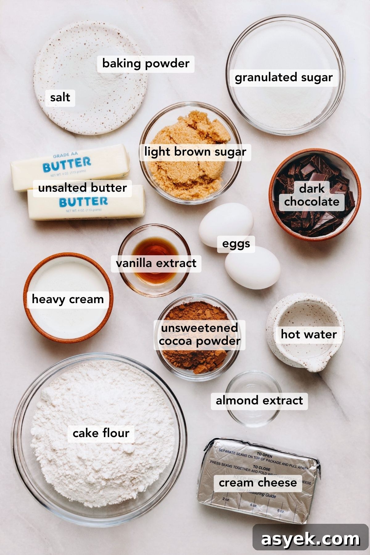 Classic Marble Bundt Cake Recipe with Chocolate Swirls 4 Ingredients in various size bowls and plates labeled as salt, baking powder, granulated sugar, light brown sugar, unsalted butter, heavy cream, vanilla extract, almond extract, cream cheese, hot water, unsweetened cocoa powder, cake flour, dark chocolate, and eggs on a white marbled background.