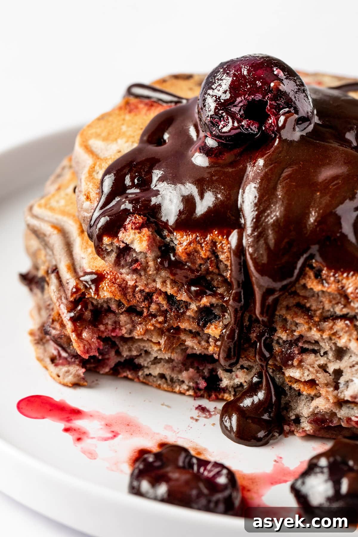 Fluffy Cherry Pancakes with Maple Syrup 7 A stack of cherry pancakes with a bite missing, topped with chocolate sauce and a fresh cherry.