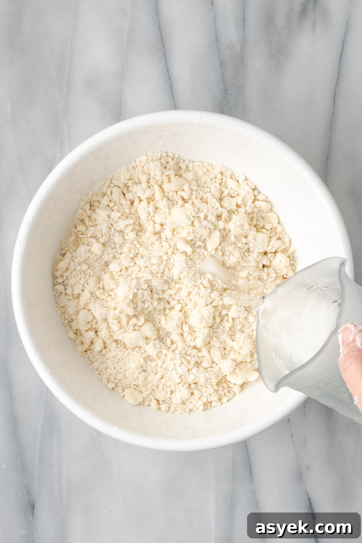 A measuring cup of water being poured over the partially mixed pie dough.