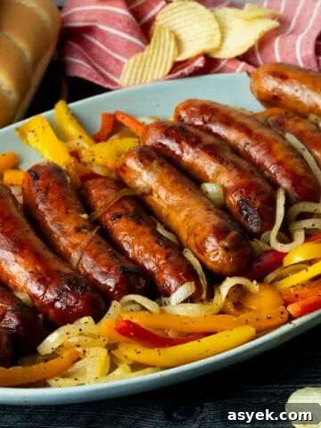 Grilled sausage atop onions and peppers on blue platter with buns and potato chips in background.