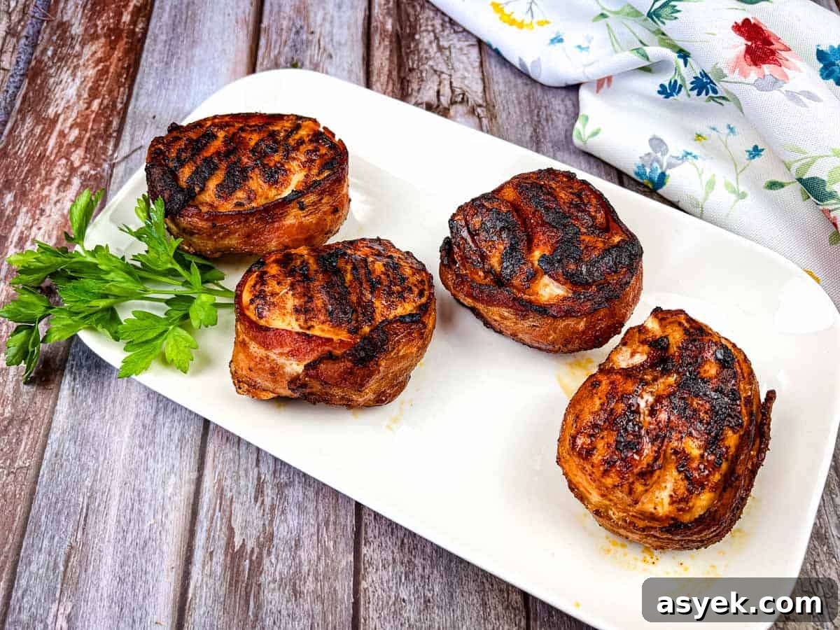 Four bacon-wrapped chicken breasts on a white rectangular plate garnished with sprigs of parsley.